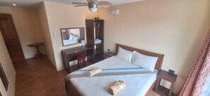 A hotel room with a bed, a desk, a chair, a mirror, a ceiling fan, and a door. - Cobadonga Inn Alona (Panglao)