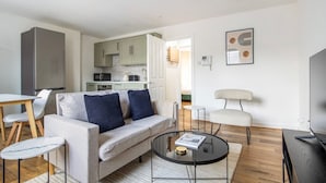 Un salon moderne avec un canapé, une table basse et un téléviseur. On aperçoit une cuisine à l’arrière-plan, avec des placards et des appareils électroménagers.