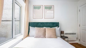 Une chambre avec un grand lit, une tête de lit verte, deux œuvres d’art encadrées, un radiateur et une fenêtre avec des rideaux.