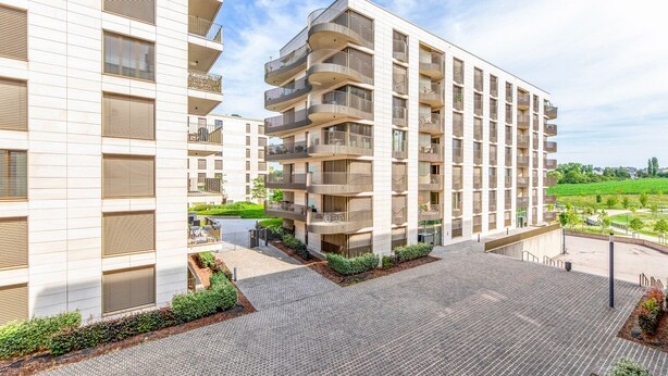 A modern apartment building with multiple balconies, a paved courtyard, and a landscaped garden area.