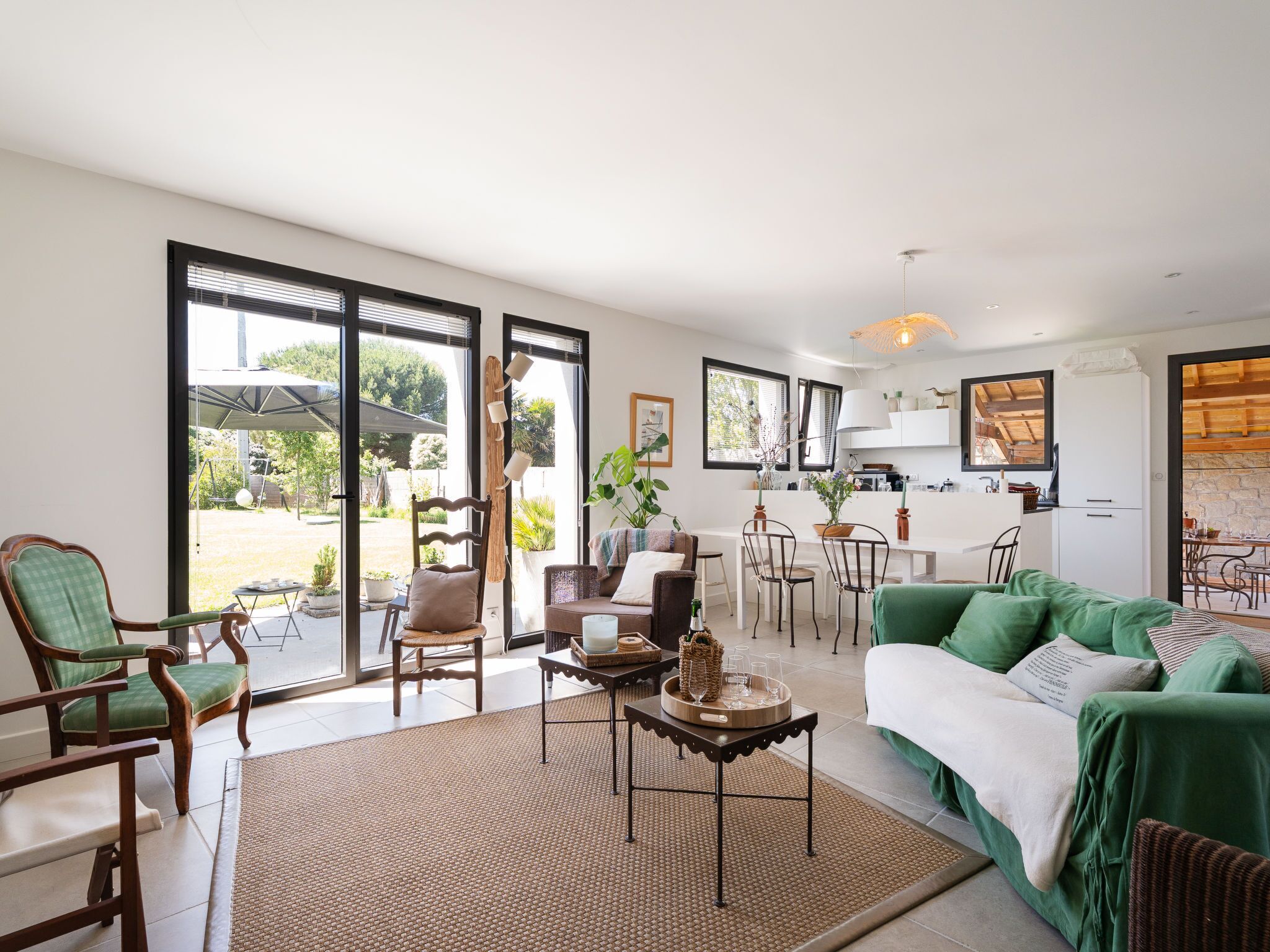 A spacious living area with a green sofa, wooden chairs, a dining table, and a kitchen in the background.