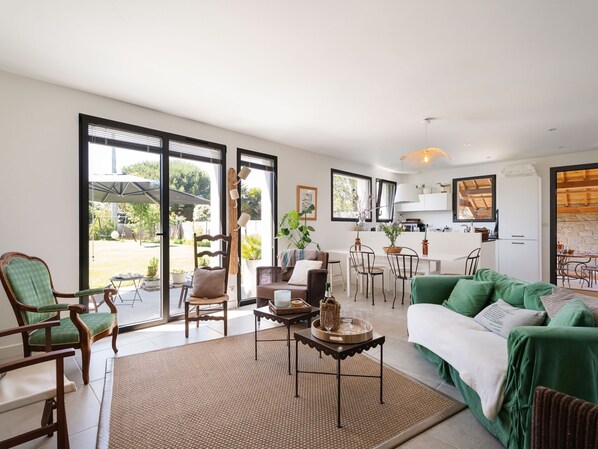 A spacious living area with a green sofa, wooden chairs, a dining table, and a kitchen in the background.