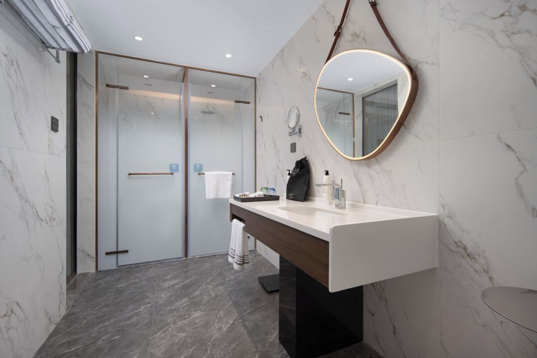 A modern bathroom with a marble floor, a large mirror with a gold frame, a white sink, and a glass shower enclosure.