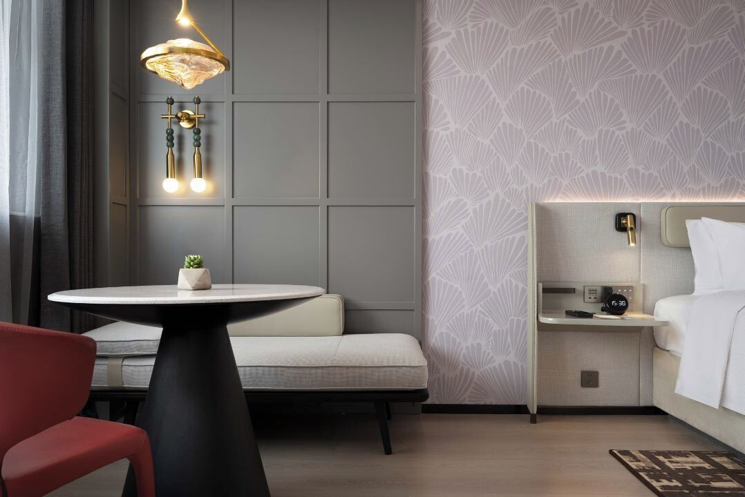 A modern hotel room with a round table, a red chair, a sofa, a bed, a nightstand, and a wall-mounted lamp.