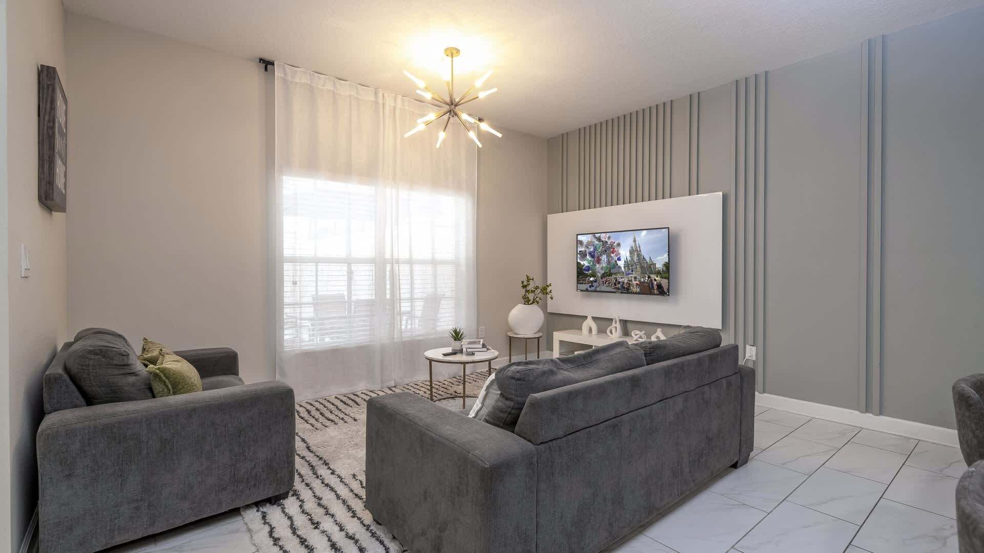 A modern living room with a grey sofa, a TV mounted on the wall, and a decorative ceiling light.