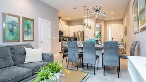 A modern living room with a grey sofa, dining area with chairs, a kitchen in the background, and a ceiling light fixture.