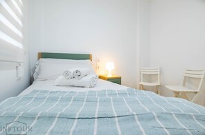 A neatly made bed with a blue striped blanket, white pillows, and rolled towels. There is a bedside lamp on a wooden nightstand. Two white chairs are against the wall. - Inftour Medici 6L Apartamento (Calp)