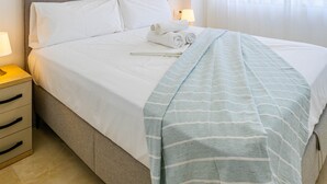 A neatly made bed with white linens, a grey headboard, and a striped blanket. There is a nightstand with a lamp and a drawer. - Inftour Medici 6L Apartamento (Calp)