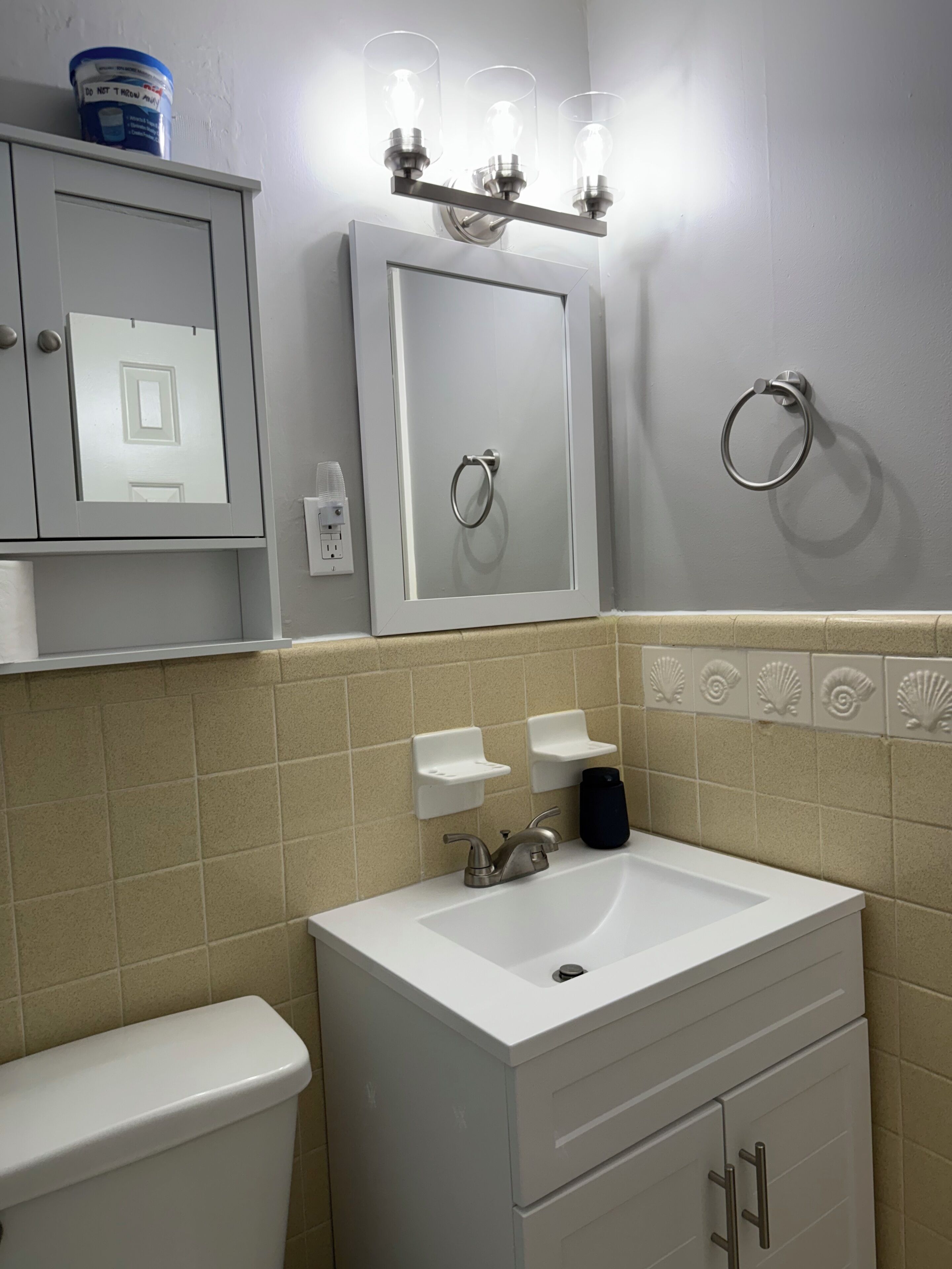 Un baño con lavamanos blanco, inodoro, espejo con argolla para toallas y estante con un recipiente azul.