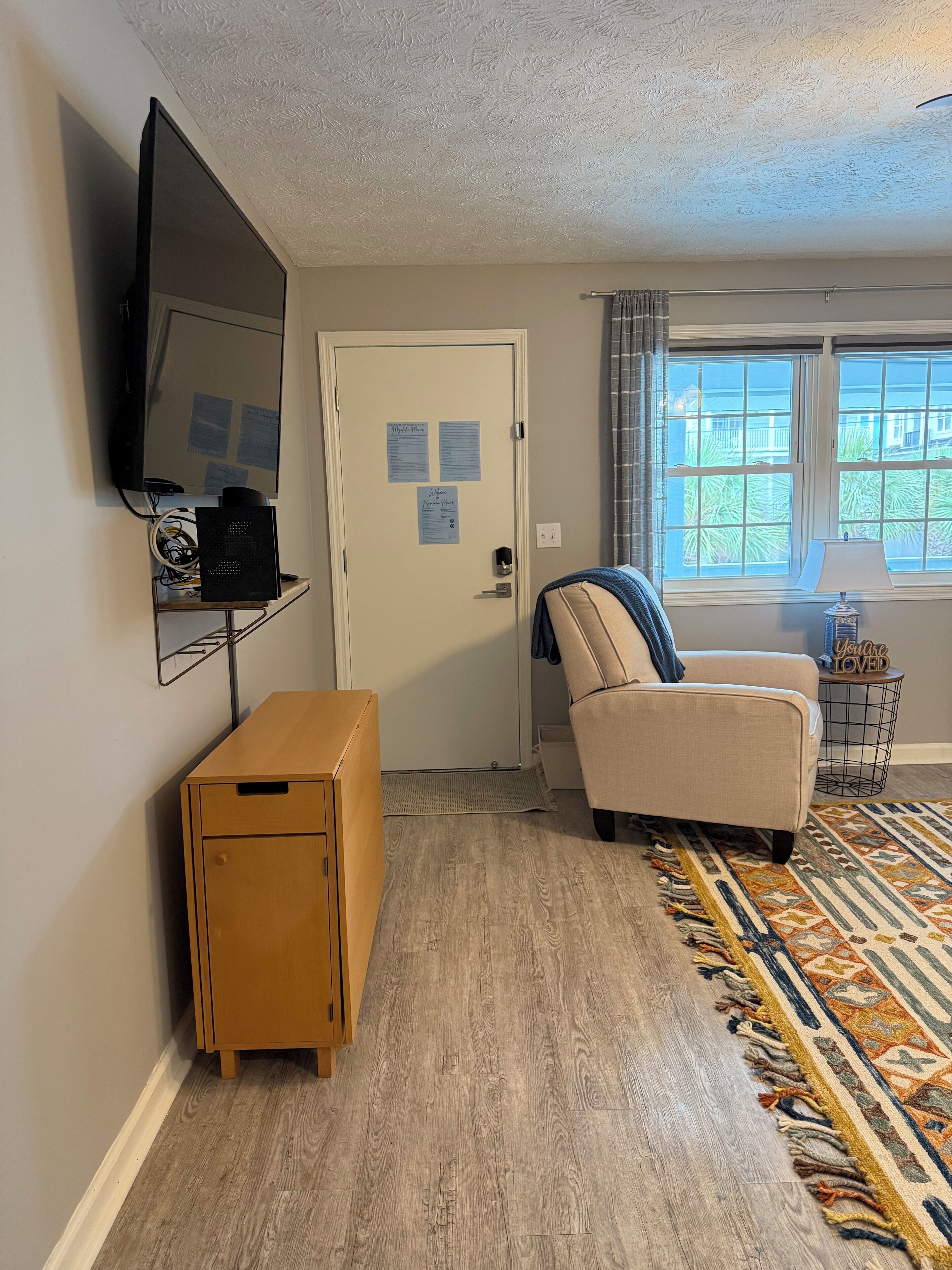 Ein Wohnzimmer mit einem Holzkasten, einem beige gefärbten Sessel, einem gemusterten Teppich, einem an der Wand befestigten Fernseher und einer Tür mit Aushängen.