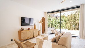 A modern living room with a sofa, a wooden cabinet, a TV, and a large window with a view of greenery. - 3Br Khana in Canggu With 3 Bedrooms and 3 5 Bathrooms (Canggu)