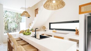 A modern kitchen with a large island, wicker chairs, and a large window providing a view of greenery. - 3Br Khana in Canggu With 3 Bedrooms and 3 5 Bathrooms (Canggu)