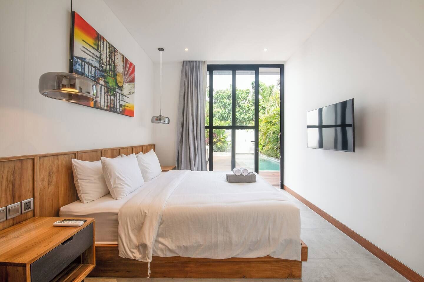 Ein modernes Schlafzimmer mit einem Holz-Kopfteil, einem großen Bett, einem Nachttisch mit Lampe, einem an der Wand montierten Fernseher und Blick auf einen Swimmingpool durch eine Glastür.