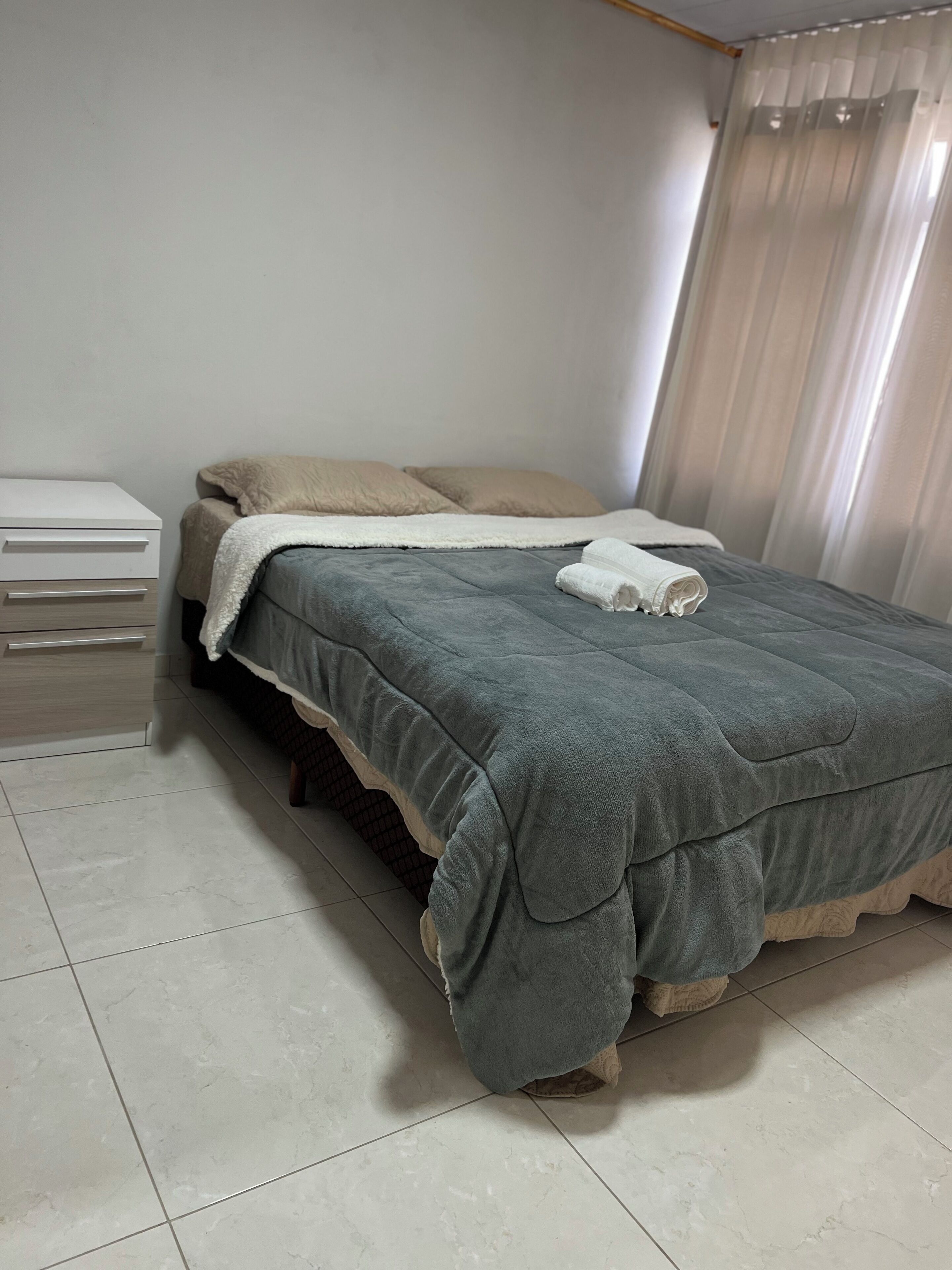 Un lit bien fait, avec une couette grise, un oreiller blanc et une serviette pliée posée sur le lit. Une table de chevet blanche, dotée de tiroirs, se trouve à gauche du lit. La pièce est dotée d’un sol carrelé et d’une fenêtre ornée de rideaux légers.