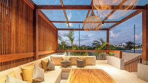 A modern outdoor seating area with a wooden bench and cushions, a glass ceiling, and a view of palm trees and buildings.