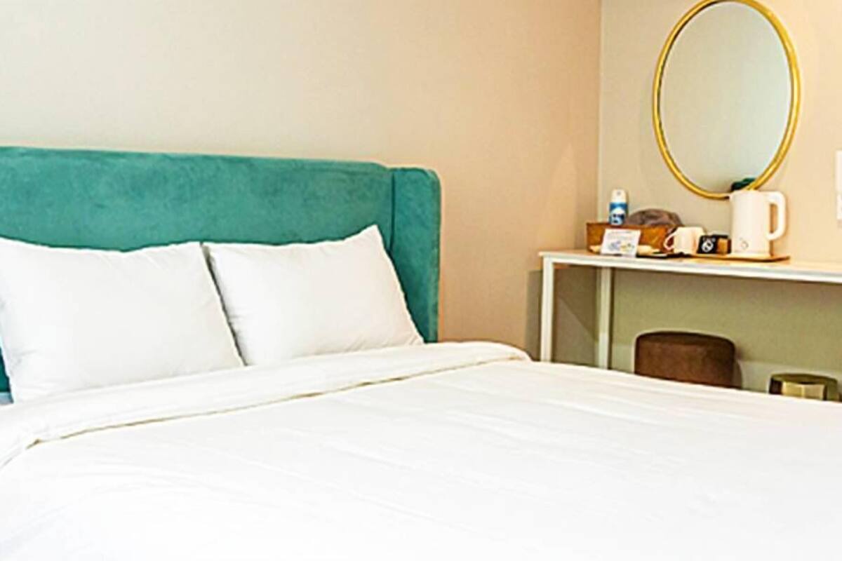 A hotel room with a teal headboard, white bedding, a round mirror, a small table with a coffee maker, and a chair.