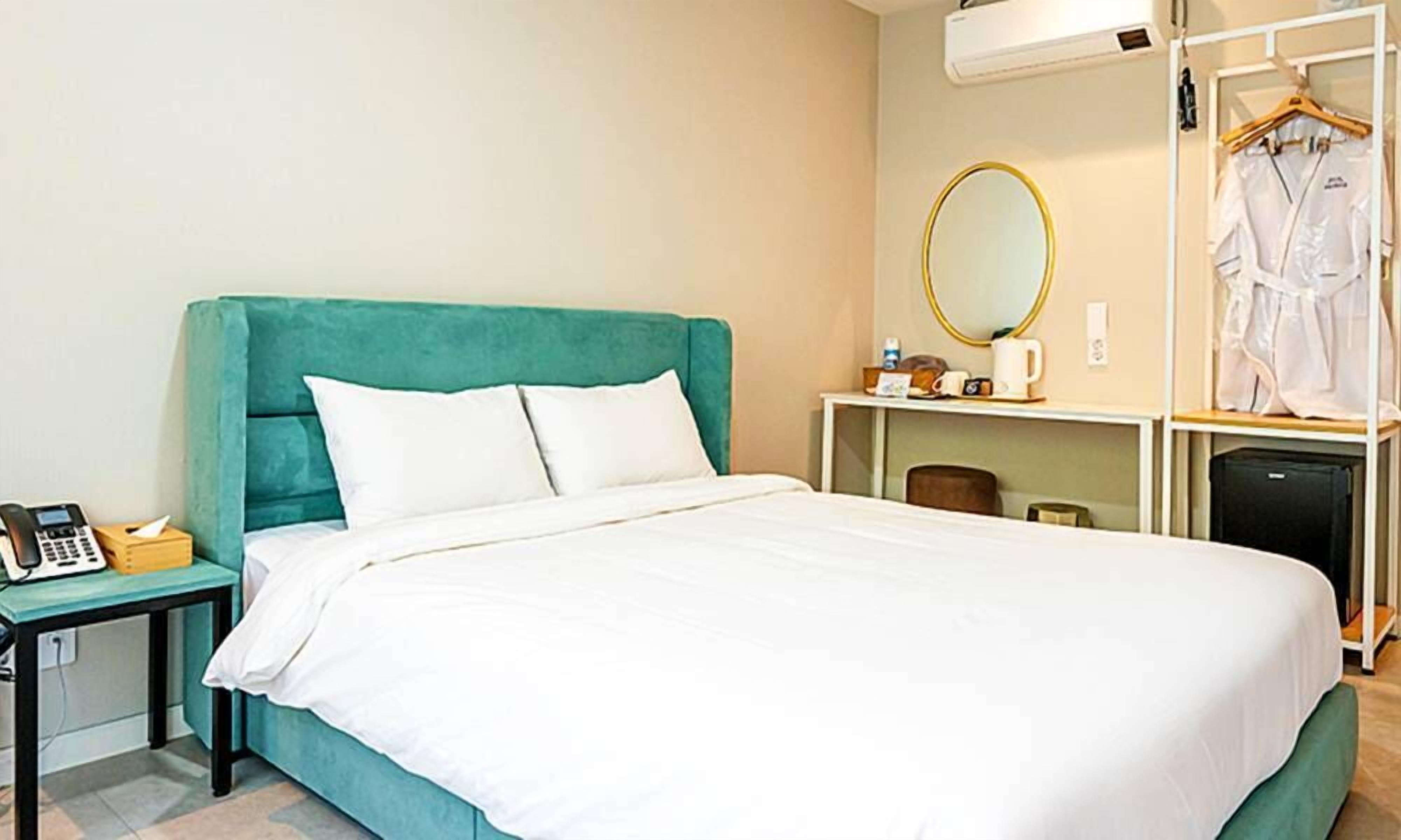A neatly made bed with white bedding, a teal headboard, a bedside table with a telephone, a small table with a cup and a tissue box, a mirror, and a clothing rack with a white garment.