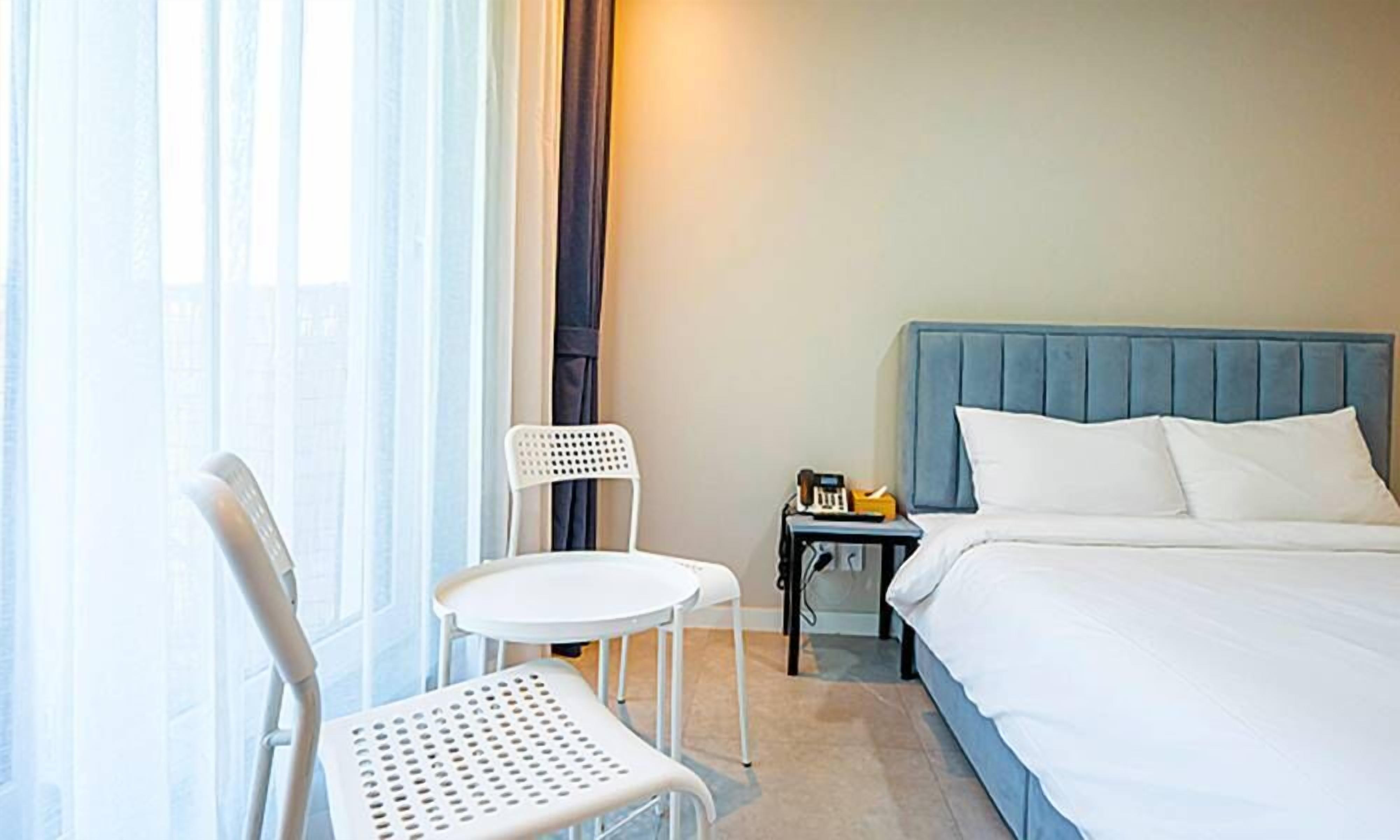 A hotel room with a bed, a chair, a small table, and a nightstand with a lamp.
