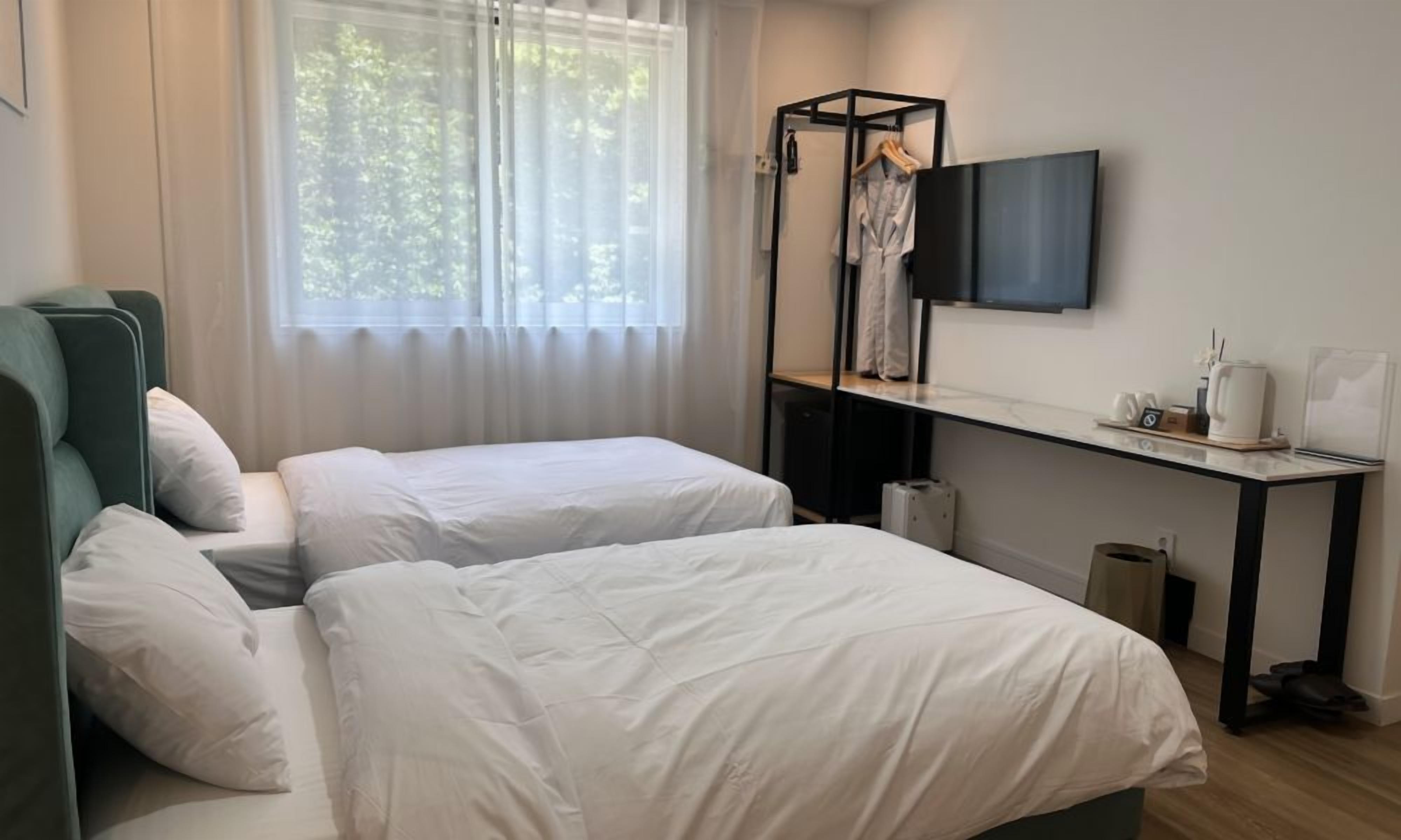 A hotel room with two beds, a desk with a flat-screen TV, and a window with sheer curtains.