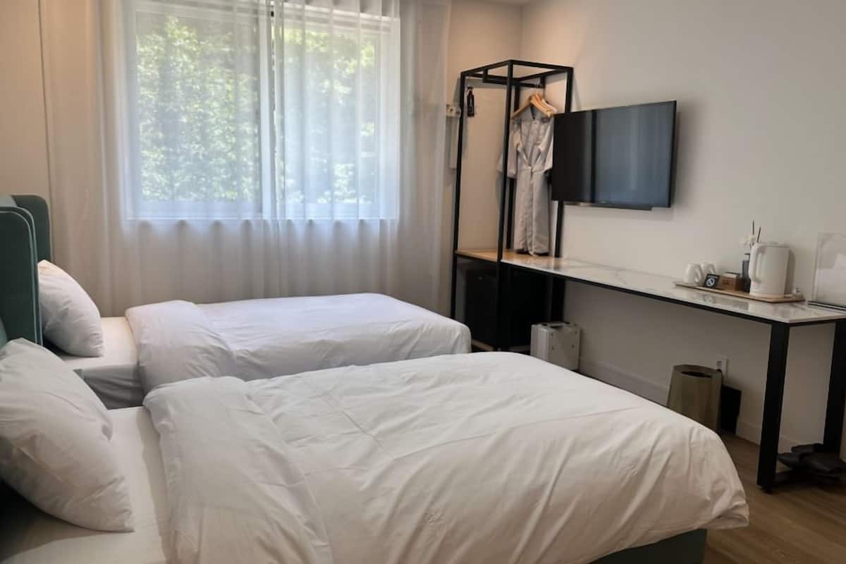 A hotel room with two beds, a desk with a flat-screen TV, and a window with sheer curtains.