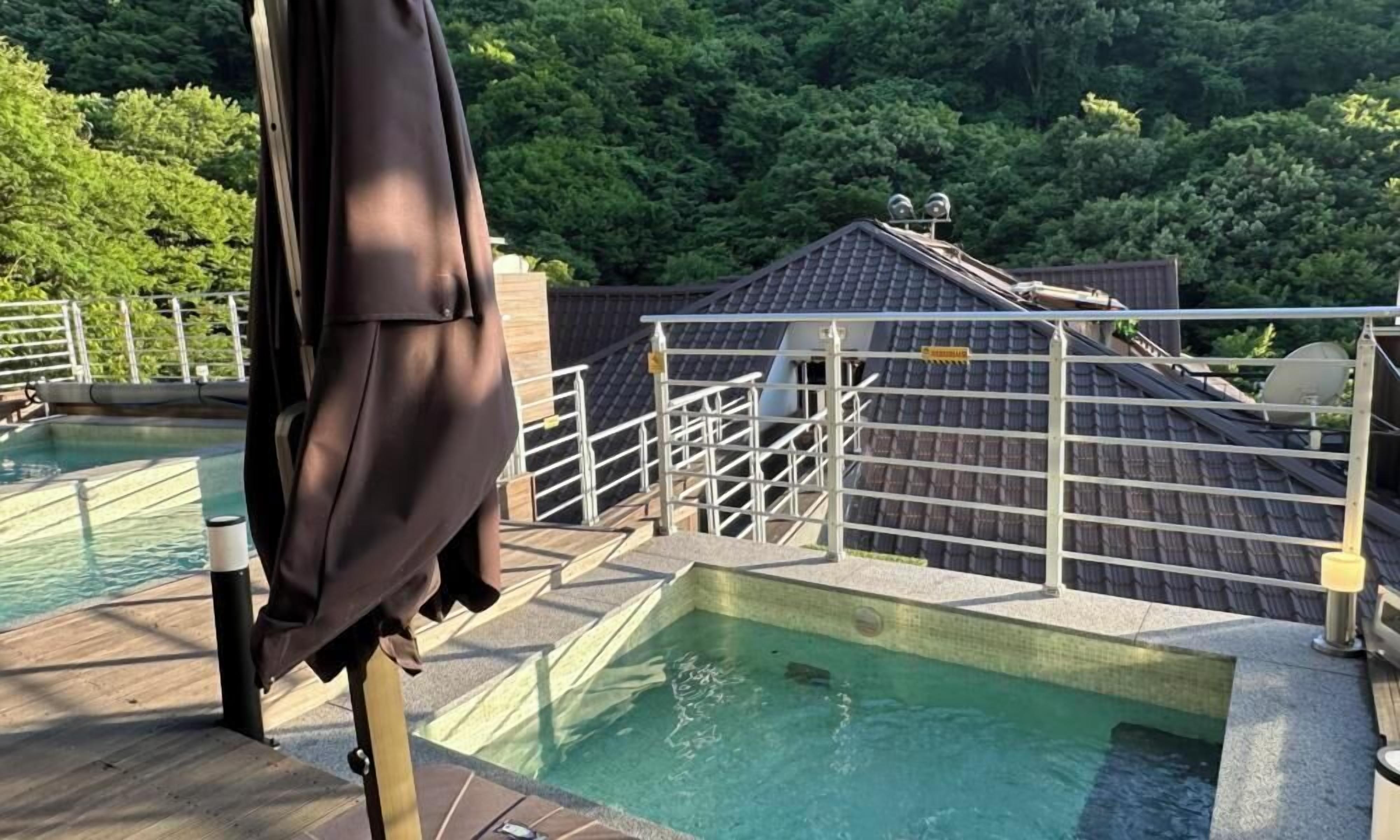 A swimming pool with a wooden deck, surrounded by a metal railing and a view of a forested area.