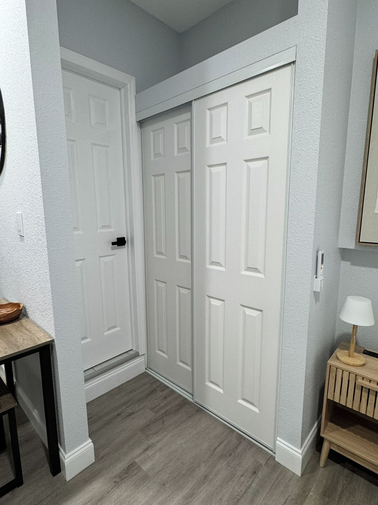 A hallway with a white door, a small table with a bowl, and a lamp on a wooden side table.