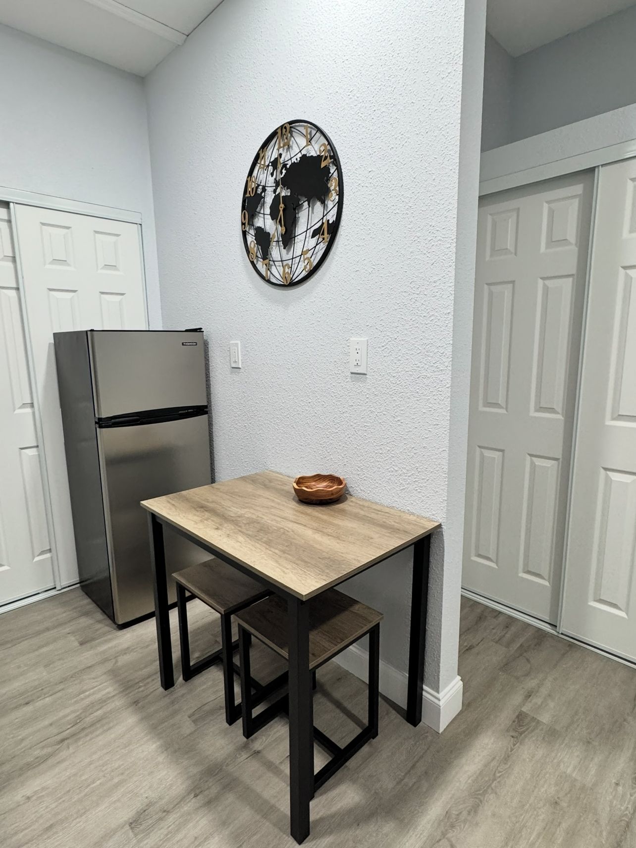 Una cocina con una mesa de madera, sillas negras, un refrigerador de acero inoxidable y un cuadro con un mapa del mundo en la pared.