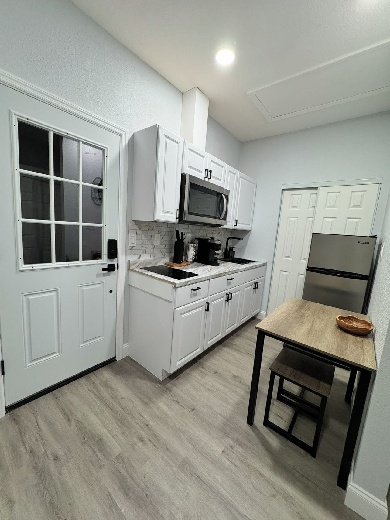 Cocina compacta con muebles blancos, microondas, refrigerador y una mesa con banco.