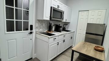 Cocina compacta con muebles blancos, microondas, refrigerador y una mesa con banco.