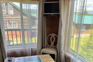 A room with a large window, a wooden chair, a table with a painting, and a television mounted on the wall. - Chenik Cabin, Homer Floatplane Lodge (Homer)