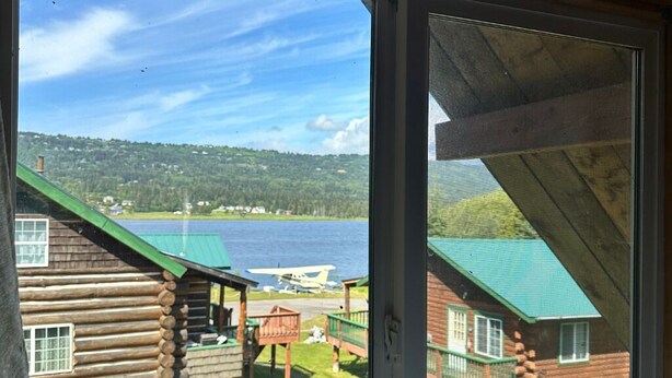 A log cabin with a green roof and a view of a lake and a small airplane in the background. - Chenik Cabin, Homer Floatplane Lodge (Homer)