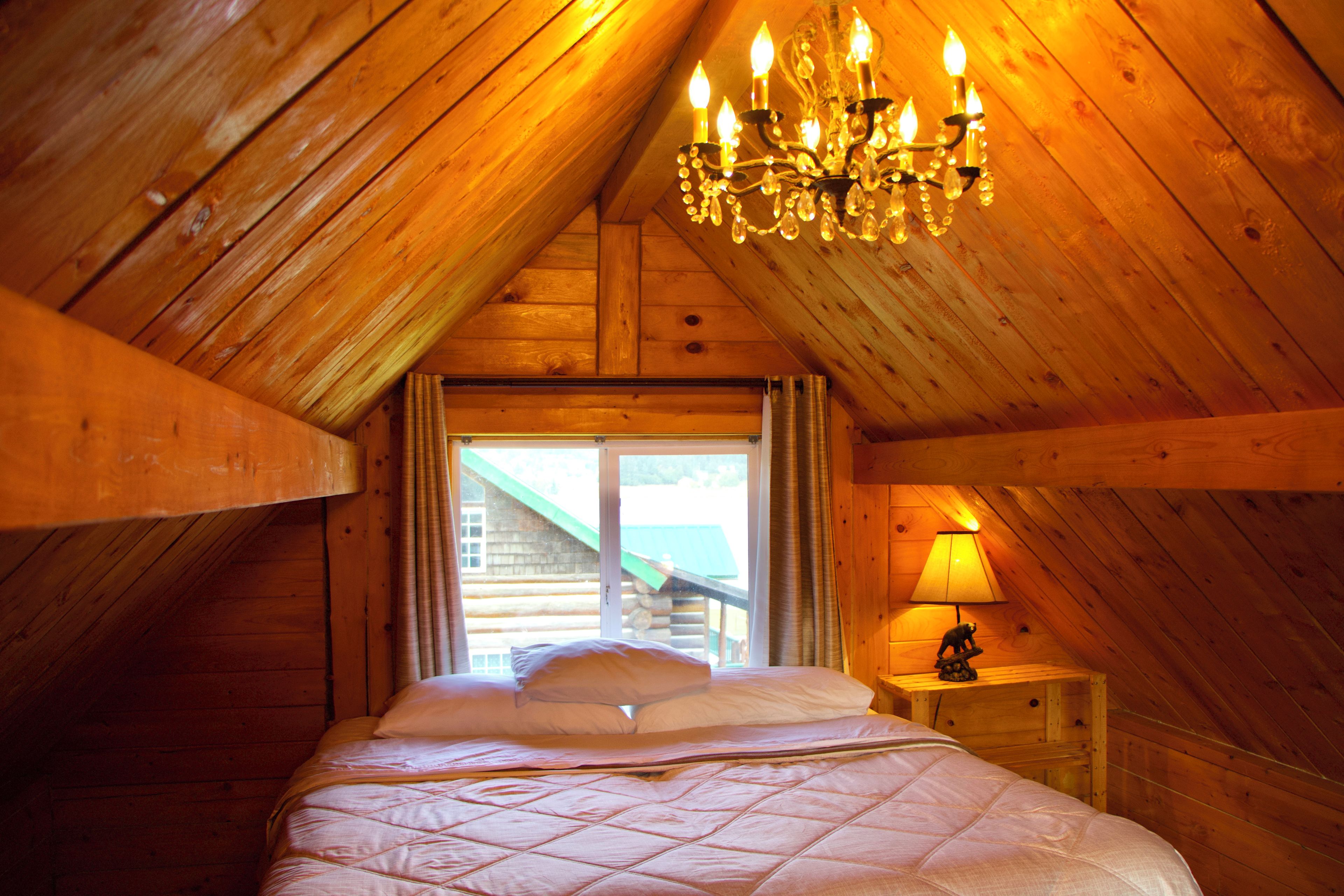 A cozy attic bedroom with a wooden sloped ceiling, a chandelier, a bed with pillows, a nightstand with a lamp, and a window with curtains.