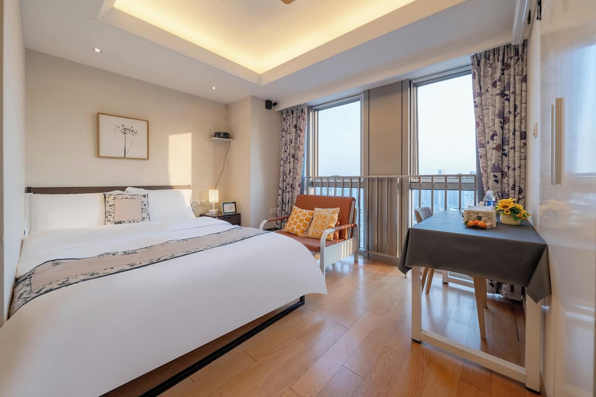 Une chambre d’hôtel avec un grand lit, une petite table sur laquelle est posé un vase de fleurs, une chaise et un balcon avec des rideaux.