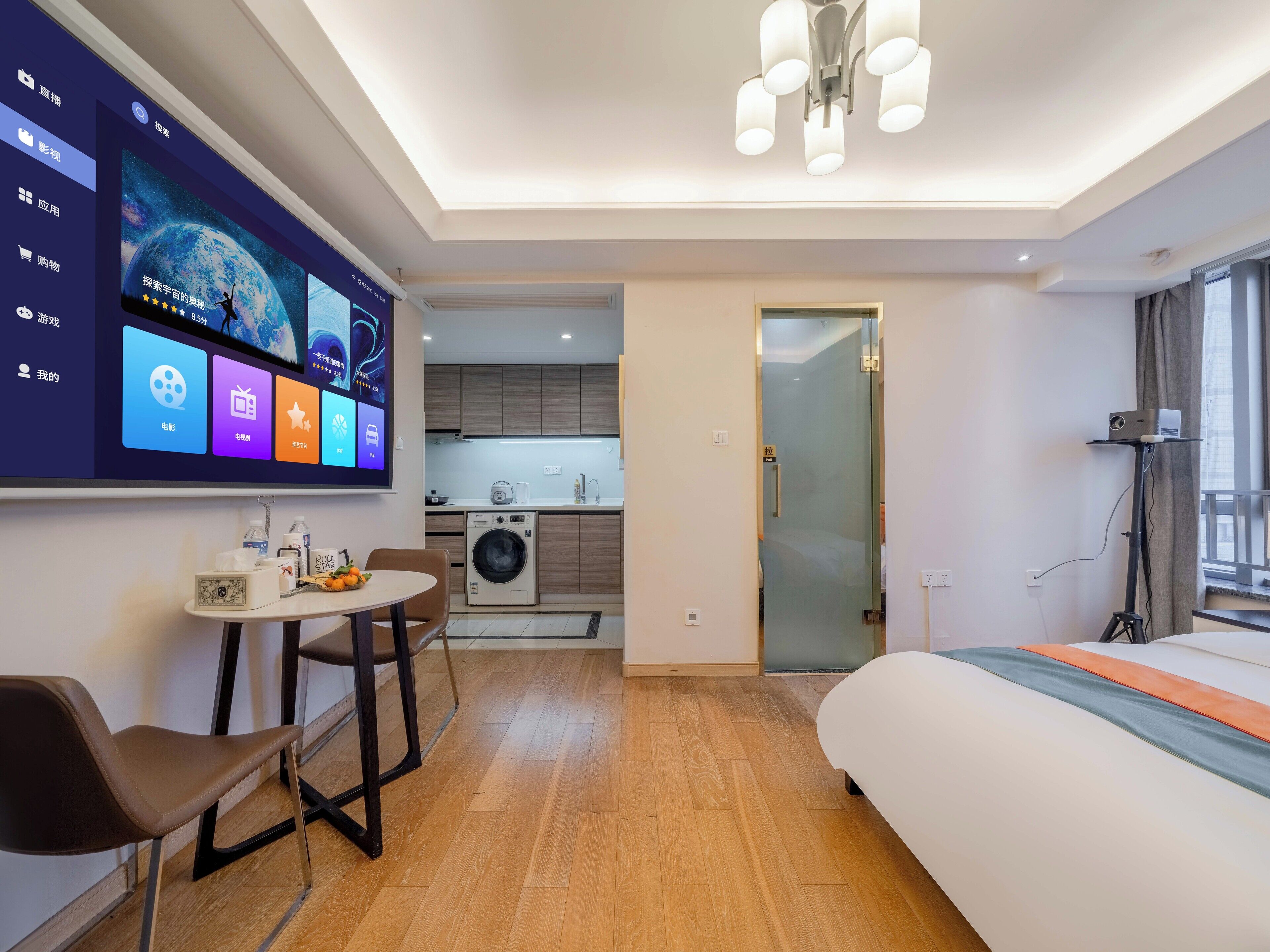 Une chambre d’hôtel moderne équipée d’un écran numérique, d’un lit, d’un bureau avec une chaise, d’un lave-linge et offrant une vue sur la cuisine et le balcon.