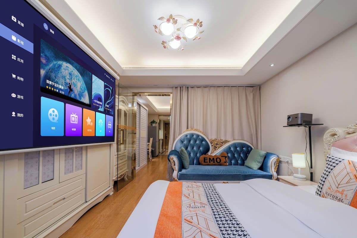 Une chambre d’hôtel moderne dotée d’un grand téléviseur à écran plat affichant une application hôtelière, d’un canapé bleu agrémenté de coussins décoratifs, d’un lit recouvert d’une couvre-lit à motifs et d’une table de chevet avec une lampe.