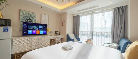 A hotel room with a large bed, a desk, a chair, a TV, and a balcony with curtains.