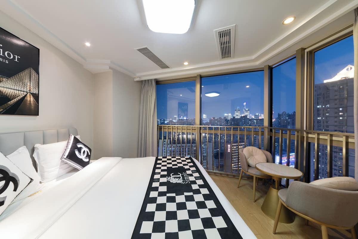 Une chambre d’hôtel dotée d’un grand lit, d’un tapis à motifs à carreaux, d’un balcon offrant une vue sur la ville et d’un coin salon avec une table et des chaises.