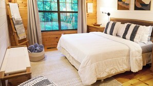 Ein Schlafzimmer einer Holzhütte mit einem großen Bett, einem Holzstuhl, einem kleinen Tisch und einem Fenster mit Blick auf Bäume.