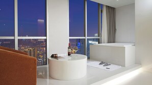 Une chambre d’hôtel moderne dotée d’une grande fenêtre donnant sur la ville, d’une table ronde blanche avec un magazine et des verres, et d’une baignoire blanche.