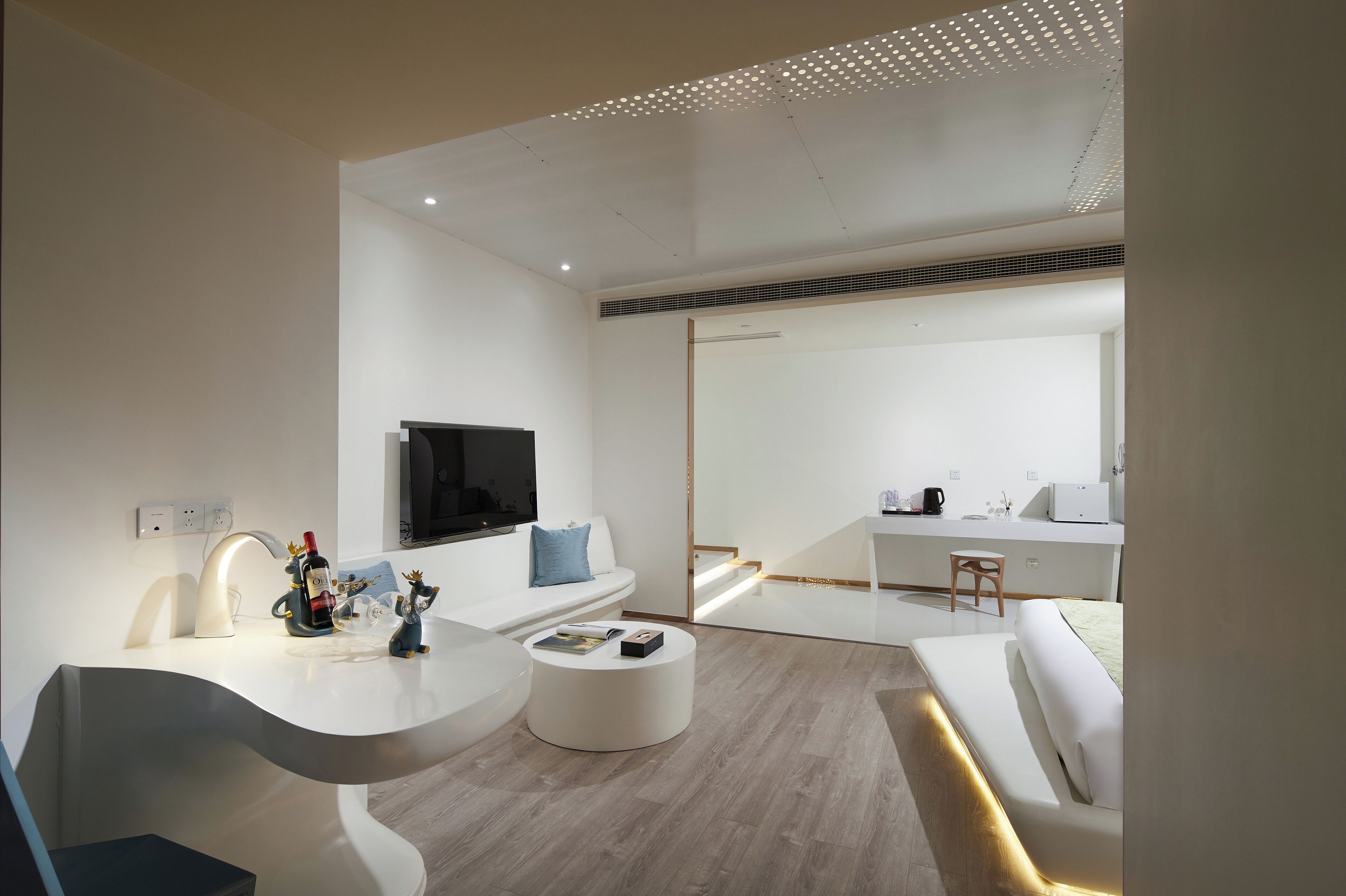 A modern, minimalist living room with a curved white console table, a white sofa, and a round coffee table. A flat-screen TV is mounted on the wall above the sofa.