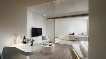 A modern, minimalist living room with a curved white console table, a white sofa, and a round coffee table. A flat-screen TV is mounted on the wall above the sofa.