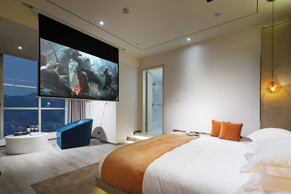 Een moderne hotelkamer met een grote televisie, een bank, een bed met oranje beddengoed en uitzicht op de stad.