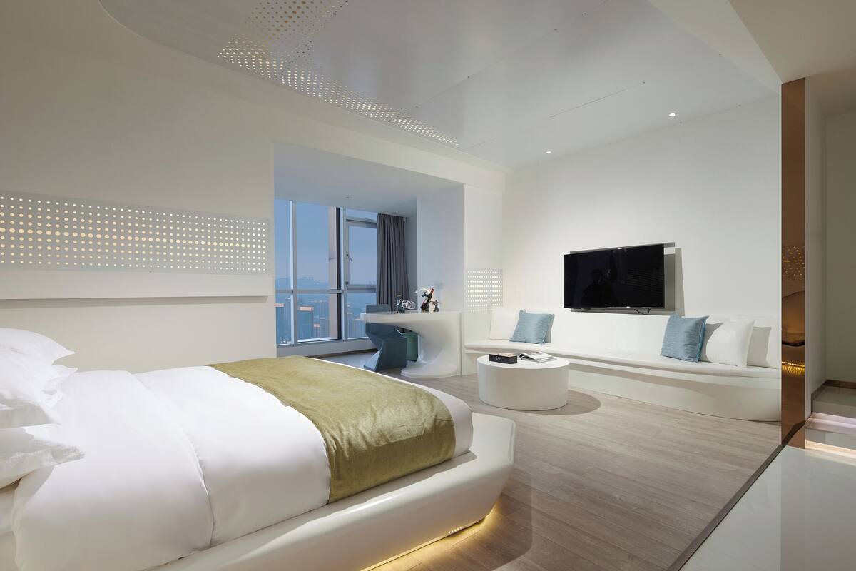 Een moderne hotelkamer met een groot bed, een zithoek met een televisie en een balkon met uitzicht op de stad.