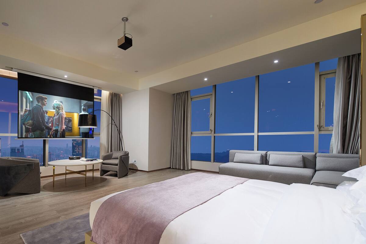 Een moderne hotelkamer met een groot bed, een bank, een salontafel en een televisiescherm waarop een film draait.