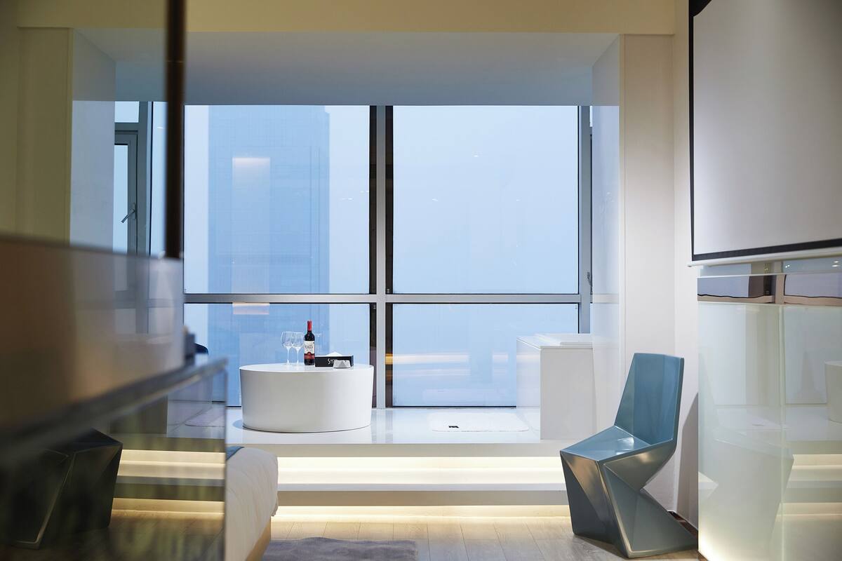Een moderne hotelkamer met een groot raam, een witte ronde tafel met een fles en glazen, een blauwe stoel en een glazen balustrade.