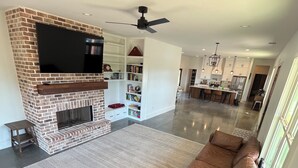 Un salon moderne avec une cheminée en briques, un grand téléviseur à écran plat, un canapé d’angle et un coin repas avec une cuisine en arrière-plan.
