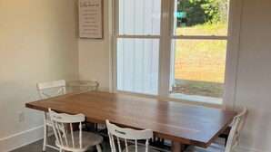 Un coin repas avec une table en bois et des chaises blanches, une fenêtre avec des stores et une citation inspirante sur le mur.