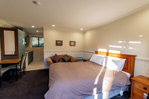 A bedroom with a large bed, a desk, and a chair. There is a window with blinds and a door to the left.