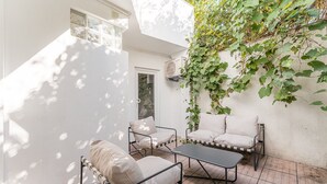 Une terrasse avec un canapé à ossature métallique, une petite table et une chaise. On y trouve un mur blanc avec une fenêtre et un appareil de climatisation.
