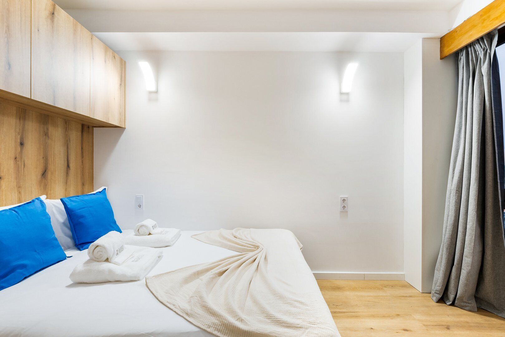 Une chambre d’hôtel moderne avec une tête de lit en bois, un lit avec du linge de lit blanc et des oreillers bleus, un plaid beige et un rideau gris.